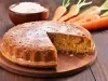 Bolo de cenoura recém-assado em uma tábua de madeira. Com uma crosta dourada e polvilhado com açúcar de confeiteiro, o bolo está parcialmente fatiado, cercado por ingredientes como farinha e cenouras.