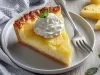 Fotografia de uma fatia grande uma torta de abacaxi com um pouco de chantilly por cima e uma folha de hortelã, sobre um prato médio raso e branco. O prato está sobre uma mesa de madeira branca.