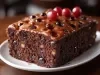 Fotografia de fatia de um bolo de chocolate com frutas, apresentando uma textura densa e escura, decorado com cerejas brilhantes e um brilho no topo, em um prato branco.
