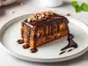 Fotografia de uma fatia de bolo com dourado e pedaços brancos, coberta com ganache de chocolate e nozes, servida em um prato cerâmico, com um toque de chocolate no fundo.