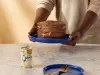 Fotografia mostra uma pessoa segurando um bolo com ganache de chocolate, com decoração rústica.