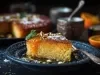 Fotografia de uma fatia de bolo laranja em um prato escuro, com calda brilhante e guarnições de açúcar de confeiteiro, pistaches e hortelã, destacando a textura e a cor quente.
