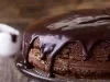 Um bolo de chocolate redondo, com textura densa e caseira, está coberto por uma espessa camada de cobertura de chocolate brilhante que escorre pelas laterais. Ele está sobre um suporte de vidro, pronto para ser servido.