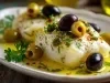 Em cima de uma mesa, prato com pedaço de bacalhau, azeitonas pretas e verdes e azeite, decorado com ervas.