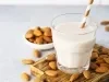 Fotografia em tons claros de um copo de vidro com um canudo dourado e branco dentro do copo, que tem uma bebida vegetal de amêndoas. O copo está sobre uma mini tábua de madeira quadrada, apoiada em uma mesa cinza clara com amêndoas espalhadas.