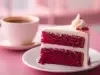 Fotografia de uma fatia de bolo veludo vermelho em um prato branco, coberta com creme branco e decorada com migalhas vermelhas, acompanhada de uma xícara de café.