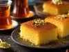Fotografia de um doce tradicional, "Revani" ou "Basbousa", em fatias quadradas com cobertura dourada e interior amarelo. Decorado com pistachios, servido com chá turco em copos elegantes.