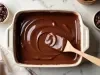 Fotografia vista de cima em tons claros de uma travessa de vidro retangular sobre uma mesa branca de mármore. Dentro do recipiente tem a sobremesa finalizada com ganache. Apoiada na travessa tem uma colher de madeira.
