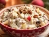 Fotografia de uma salada natalina com nozes em uma tigela vermelha decorada, destacando frutas, uvas-passas e nozes pecans envoltos em um molho cremoso.
