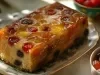 Fotografia de um bolo de frutas retangular, com cobertura caramelizada e decorado com frutas cristalizadas coloridas, apresentado em um prato oval de porcelana com detalhes florais.