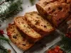 Fotografia de um bolo de frutas cortado, exibindo sua textura densa e rica, com pedaços de frutas secas e nozes. O bolo está disposto sobre uma superfície rústica, cercado por decorações de ramos verdes e bagas vermelhas.
