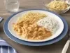 Fotografia em tons de azul em uma bancada de madeira com um sousplat azul, um prato branco raso em cima com o strogonoff, arroz branco e batata palha. Ao lado, um pano azul com listras brancas, talheres, copo com água e um potinho com batata palha.