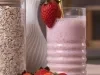 Smoothie pós-treino de morango em copo de vidro com canudo dourado e morango na borda, acompanhada por morangos frescos e pote de aveia.