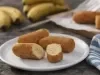 Imagem de um prato branco uma uma banana a milanesa cortada ao meio. Ao fundo, bananas e um paninho listrado branco com azul