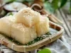 Fotografia de pedaços de bolo de tapioca quadrados dispostos em uma bandeja de bambu forrada com folha de bananeira. O coco ralado no topo e o fundo rústico.