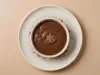 Fotografia vista de cima de um recipiente de vidro branco fundo médio com a sobremesa, finalizada com a ganache de chocolate, sobre um prato raso de vidro, com cacau em pó salpicado. O prato está sobre uma mesa de tom pastel.