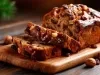 Fotografia de um bolo de frutas e nozes, com crosta dourada e textura rica. Cortado em fatias, revela um interior úmido e denso, repleto de passas e nozes, sobre uma tábua de madeira, rodeado por avelãs e passas.