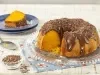 Imagem de um bolo de cenoura redondo cortado com furo central e cobertura de brigadeiro. Ao fundo, um prato azul com uma fatia de bolo e um potinho com raspas de chocolate ao lado.