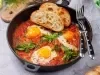 Fotografia em tons claros de uma receita de Shakshuka com pães por cima, dentro de uma frigideira de ferro preta sobre uma mesa cinza. Tem um par de talheres apoiados na frigideira, que está sobre uma mesa cinza.