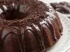 Fotografia de um bolo de chocolate em forma de bundt, coberto com um brilho de chocolate que escorre pelas laterais. Decorado com raspas de chocolate, está servido em um prato branco, com um fundo desfocado de pedaços de chocolate escuro.