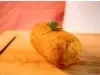 Fotografia de um croquete frito de carne moída sobre uma tábua de madeira. Tem um pequeno ramo de salsinha decorando o croquete.