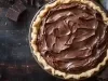 Fotografia vista de cima em tons escuros de uma torta com cobertura de chocolate sobre um praot grande e raso, apoiado em uma mesa de madeira escura.