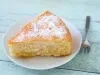 Fotografia de uma fatia de bolo de coco em um prato decorativo, com cobertura de açúcar de confeiteiro e uma crosta dourada, sobre uma superfície de madeira azul clara, acompanhada de um garfo metálico.