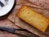 Um bolo de mandioca dourado e úmido, em formato retangular, está colocado sobre um papel manteiga. Ao lado, há uma faca rústica de madeira e um prato branco com garfos. O bolo tem aparência caseira e textura levemente crocante nas bordas.