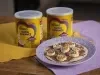 Fotografia de dois pano, roxo e amarelo, em uma mesa de madeira. Sobre esses panos tem duas latas de NINHO Nutri Advance ao lado de um prato roxo claro com uma mini pizza com chocolate e banana.