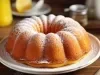 Fotografia de um bolo bundt elegantemente disposto em um prato branco, com uma leve camada de açúcar de confeiteiro. O fundo apresenta um jarro amarelo e um limão, sugerindo um ambiente acolhedor e caseiro.