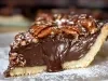 Fotografia em tons claros de uma fatia generosa de torta de chocolate com nozes sobre um prato grande e raso. Por cima tem açúcar de confeiteiro polvilhado sobre a torta e o prato.