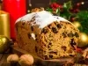 Fotografia de um bolo de frutas festivo, exibido sobre uma superfície de madeira, com textura densa e decorado com açúcar em pó. Ao redor, velas acesas, nozes e canela.