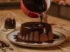 Fotografia em tons de vermelho em uma mesa de madeira com um prato branco com o bolo de chocolate em cima e uma calda de chocolate por cima. Uma panela vermelha em cima do bolo derramando a calda de chocolate.