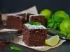 Pedaço de bolo de chocolate é o destaque, polvilhado com açúcar de confeiteiro e decorado com uma folha de hortelã. Ao lado, uma fatia de limão e mais bolos ao fundo, criando uma apresentação rústica e apetitosa.