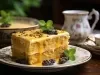 Fotografia de uma fatia de bolo de maracujá em um prato branco. O bolo, de duas camadas, possui um recheio cremoso e é coberto com um vibrante molho de maracujá, com sementes visíveis, destacando-se em um fundo suavemente desfocado.