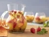 Imagem de uma jarra transparente com ponche e pedaços de frutas. Ao lado, morangos e folhas de hortelã e ao fundo dois copos da bebida.