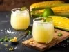 Fotografia de um copo de vidro sobre uma mini tábua de madeira retangular, sobre uma mesa preta. Ao fundo tem outro copo e três espigas de milho. Dentro dos copos tem o suco de milho, menos cremoso.