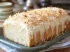Fotografia de um bolo loaf fofinho e recém-assado coberto com cobertura branca cremosa e flocos de coco tostados. O bolo está em um prato cerâmico verde, com um ambiente acolhedor ao fundo.