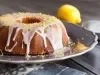 Fotografia de um bolo de limão em uma elegante travessa, coberto com glacê branco que escorre pelas laterais. Raspas de limão decoram o topo, enquanto um limão inteiro e um ralador estão ao fundo.