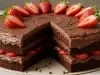Sobre uma mesa, prato com bolo de chocolate sem uma fatia. A massa é de chocolate, recheada com chocolates e morangos picados, coberta com chocolate e decorada com morangos cortados ao meio.