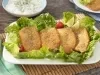Fotografia em tons de verde em uma bancada de madeira, um pano verde com azul, um jogo americano de madeira clara, um prato branco redondo com a sardinha frita acompanhada de salada de alface e tomate cereja. Ao fundo, um potinho com azeite.