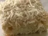 Pedaço de bolo gelado de coco coberto com coco ralado, sobre papel-alumínio em superfície clara.