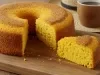 Fotografia de um bolo de milho recém-assado, com textura macia e esponjosa. Apresenta uma crosta dourada e um furo no centro, servido em uma tábua de madeira, ao lado de uma xícara de café.