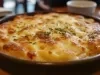 Em cima de uma mesa, refratário redondo com batata e queijo gratinado crocante, decorado com cheiro-verde picado.