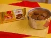 Cookie gigante em lata ao lado da embalagem Garoto Recheados Baton Creme, sobre toalhas vermelha e amarela.