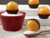 Em cima de uma mesa, pote vermelho com coco ralado, ao lado, balas redondas de caramelo crocante em forminhas marrons.