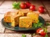 Fotografia de fatias de um bolo salgado em um prato, recheado com vegetais como milho e ervilhas. Tomates frescos e ervas verdes decoram a apresentação.