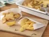 Imagem de dois saquinhos brancos de papel com batatas fritas rústicas dentro sobre uma tábua de madeira. Ao fundo, uma travessa branca com mais batatas e um pegador.