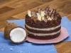 Foto da receita de Bolo Naked de Prestígio, decorado com pedaços de chocolate, sobre um prato rosa, numa bancada de madeira com tecidos azuis e dois pedaços de coco fresco aberto