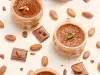 Fotografia em tons claros de quatro recipientes de vidro pequenos com a sobremesa, finalizados com a ganache, chocolate em pó, folhas de hortelã, e algumas castanhas. Os recipientes estão sobre uma mesa de tom claro, com castanhas e cacau espalhados